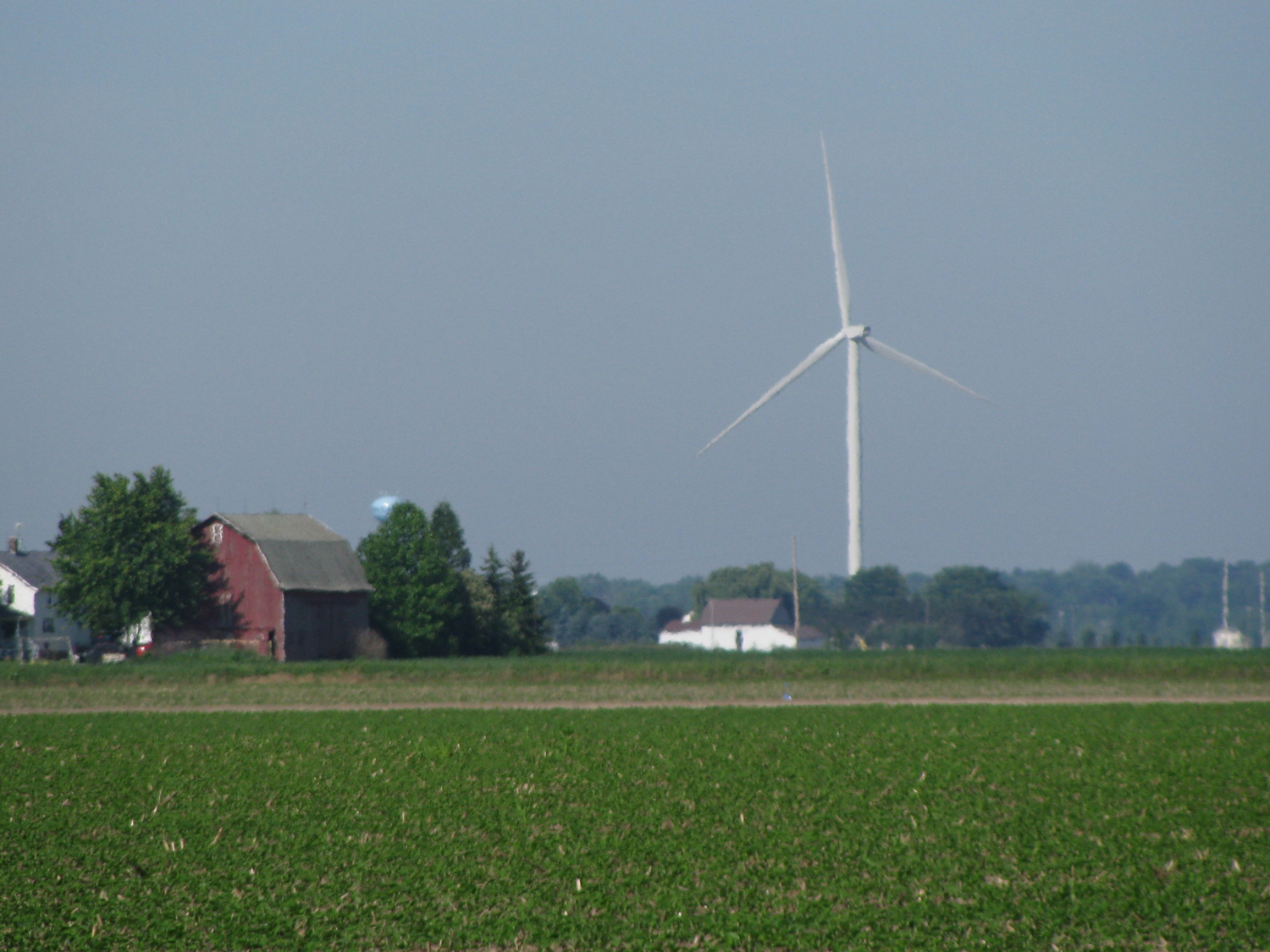 A red barn and wind turbine in the background surrounded by lush green farmland under a clear blue sky.