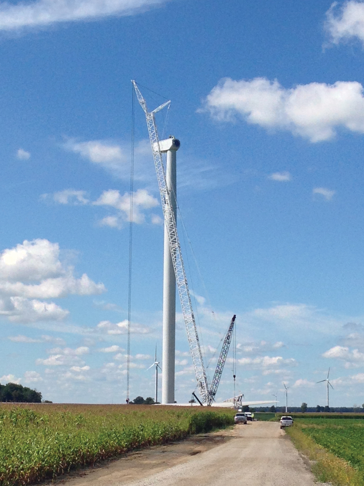 Construction crane lifting a wind turbine tower segment, with turbines and farmland in the background under a sunny blue sky.