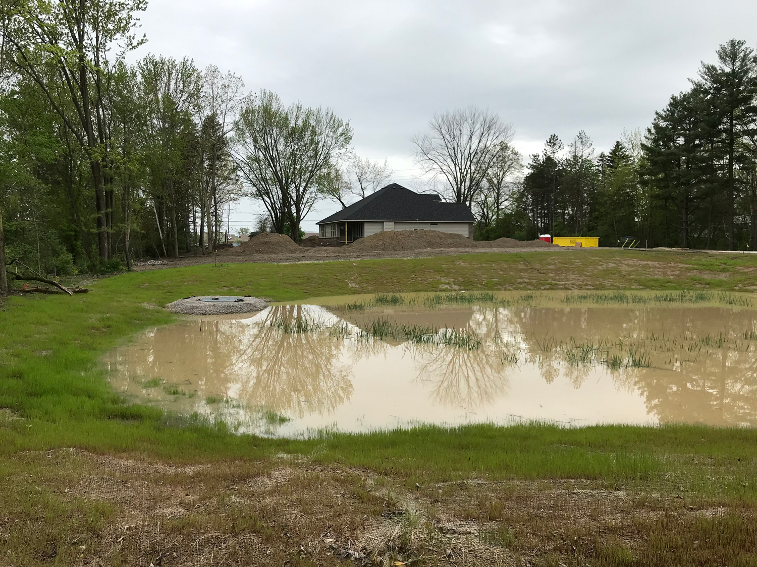 Stormwater detention pond with clear water reflecting nearby trees and a residential house under construction in the background, surrounded by green grass.