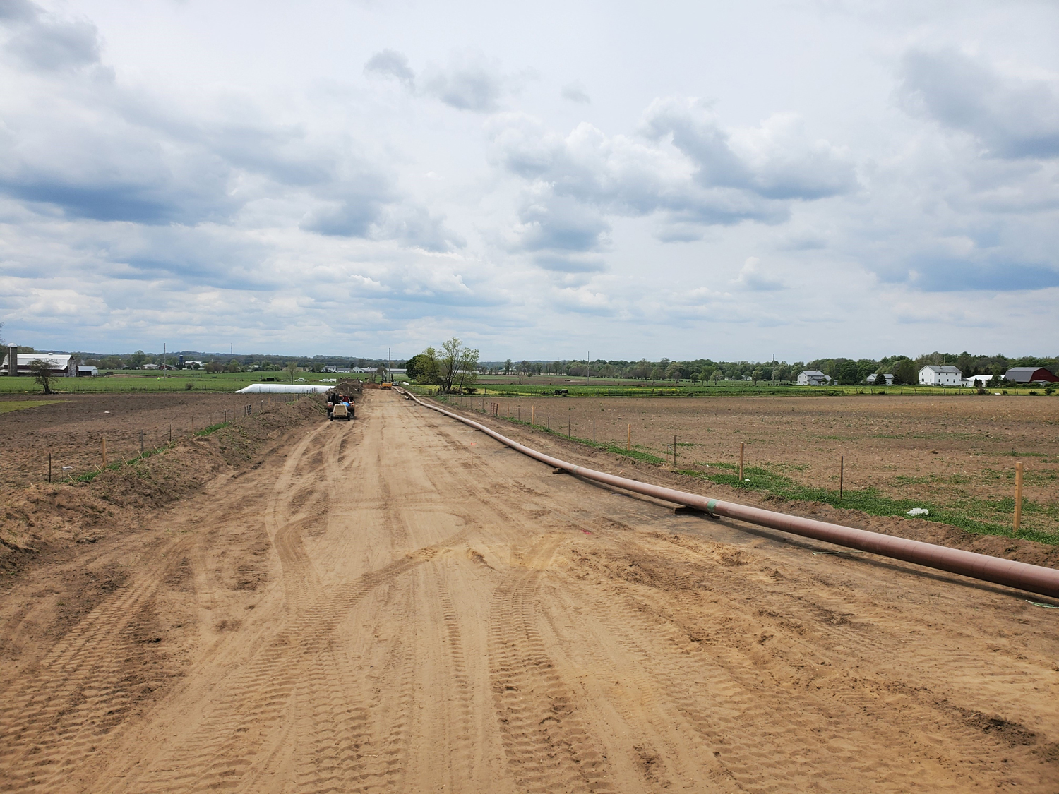 A rural construction site with a gas pipeline being installed on a dirt road, surrounded by farmland and a cloudy sky in Shipshewana, Indiana.