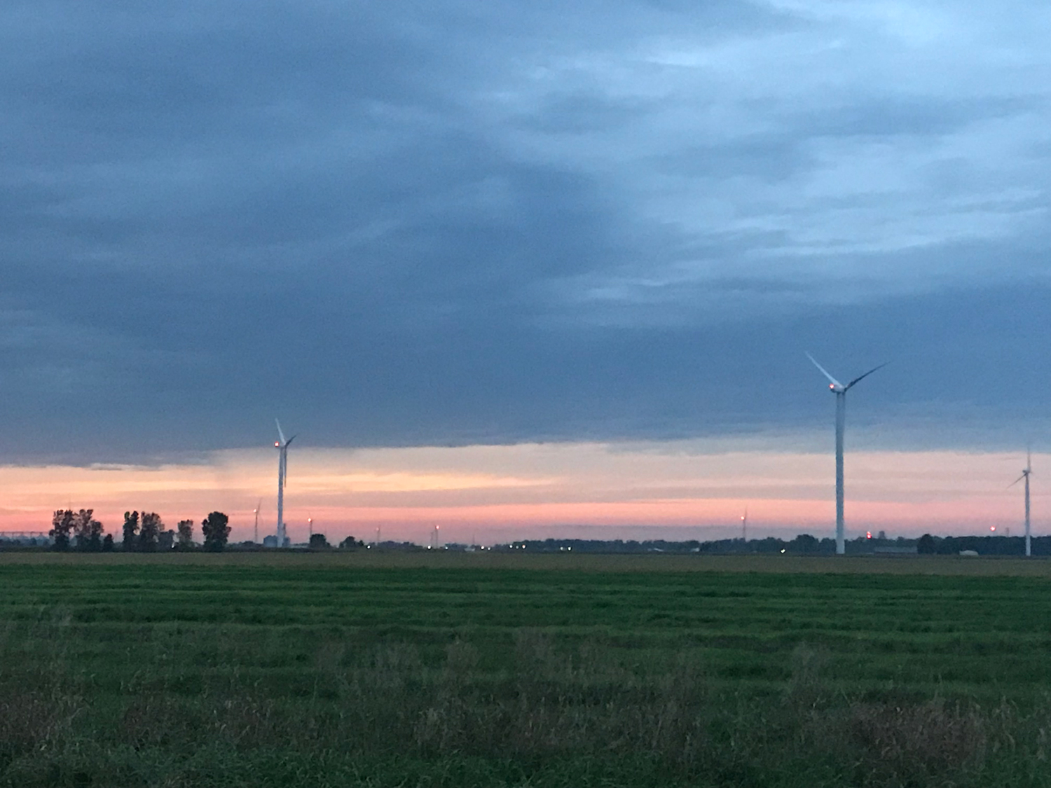 Multiple wind turbines standing in a field under a dusk sky with lights on the turbines.