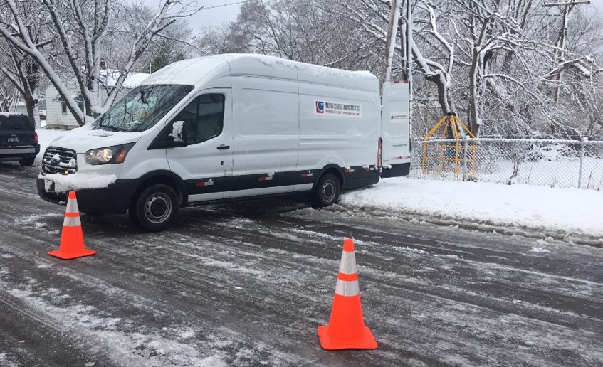 Metro Consulting Associates van parked on a snowy residential street, marked with traffic cones for safety, with manhole scanning equipment in use nearby.