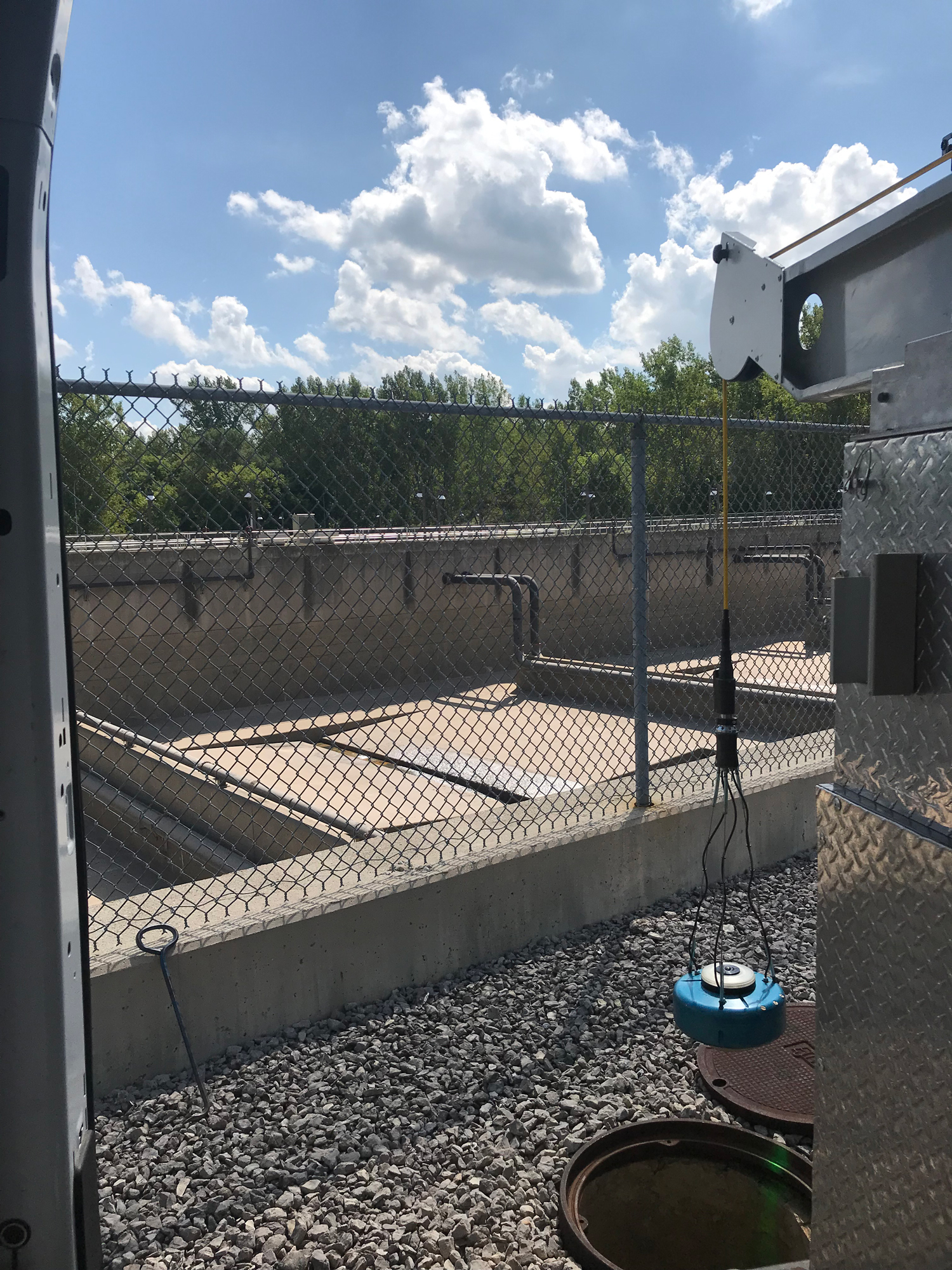 Close-up of a wastewater treatment system with a manhole scanner and equipment, surrounded by a gravel work area and fenced boundary.