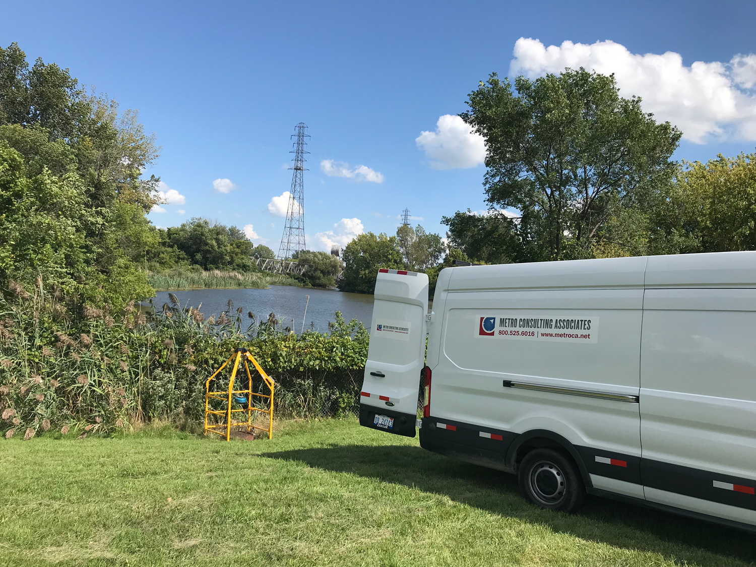 Metro Consulting Associates vans parked near a scenic pond with lush greenery and a transmission tower in the background, highlighting a field location for wastewater asset management.